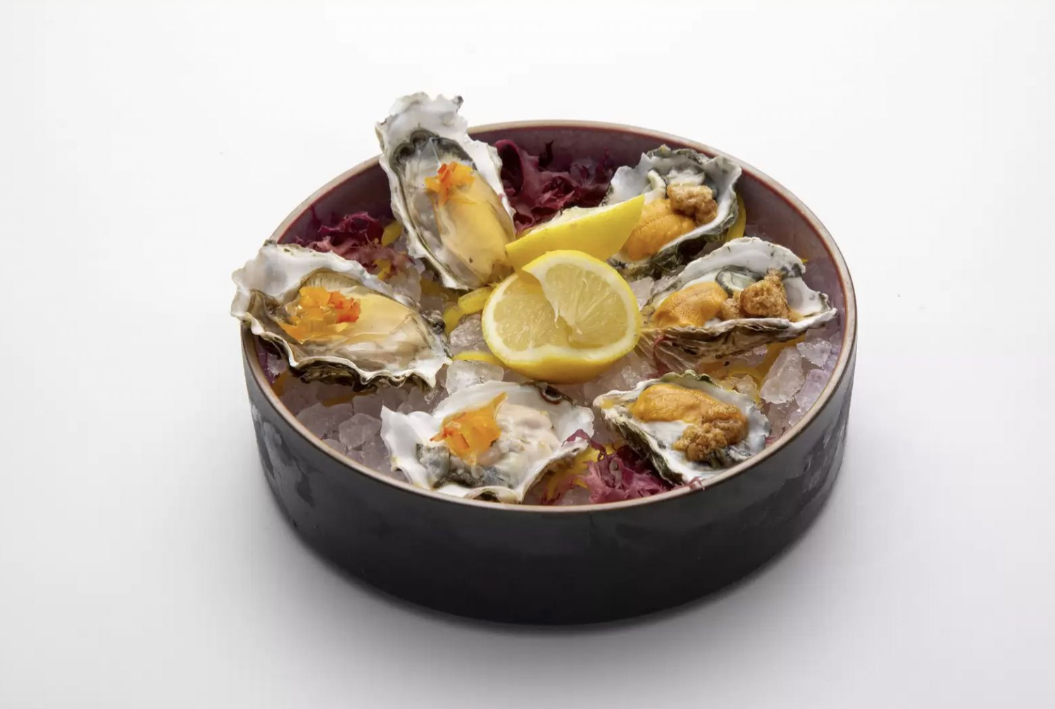 Oysters on a bed of ice with lemon wedges in a dark dish on a light background