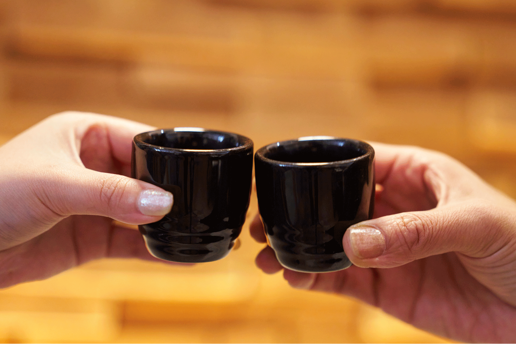 Two hands holding black ceramic cups against a wooden background